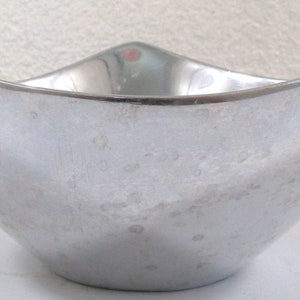 Puede incluir: Un cuenco de metal plateado con un dise&ntilde;o curvo &uacute;nico. El cuenco tiene una superficie lisa y brillante con un patr&oacute;n texturizado sutil.