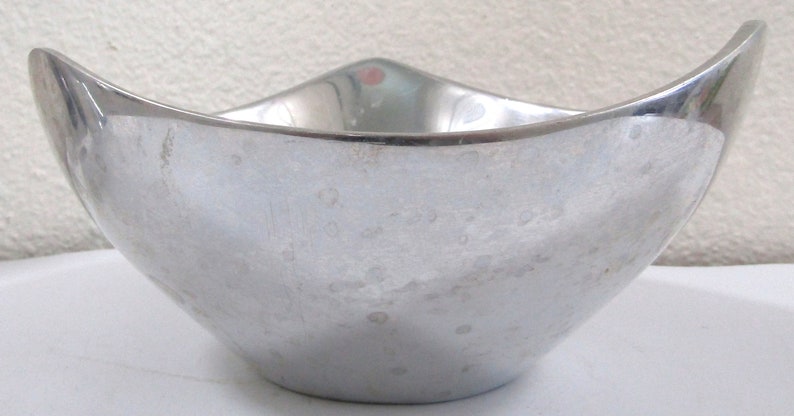 Puede incluir: Un cuenco de metal plateado con un dise&ntilde;o curvo &uacute;nico. El cuenco tiene una superficie lisa y brillante con un patr&oacute;n texturizado sutil.