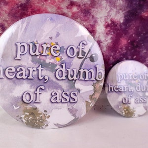 Puede incluir: Dos botones redondos con el texto "pure of heart, dumb of ass" en blanco y negrita. Los botones tienen un diseño floral de acuarela en tonos morados y verdes.