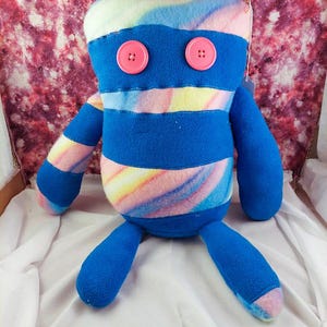 Puede incluir: Un peluche hecho a mano con forma de criatura con tela a rayas azules y arcoíris. El juguete tiene brazos y piernas azules y ojos de botón rosas. El fondo es una mezcla de rosa y rojo.