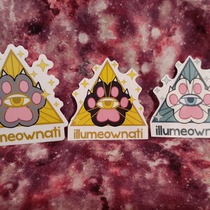 May include: Three stickers featuring a cat paw design inside a yellow triangle. The paw prints are pink, grey, and black, with an eye in the centre. The text "illumeownati" is at the bottom of each sticker.