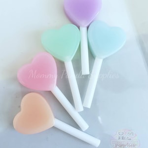 May include: Five pastel-colored heart-shaped lollipops on white sticks. The lollipops are arranged in a semi-circle, with the purple lollipop at the top and the orange lollipop at the bottom. The lollipops are all different shades of pastel pink, blue, green, purple, and orange.