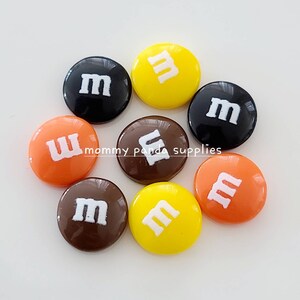 Può includere: Assortimento di bottoni di caramelle rotondi e colorati con la lettera "M" stampata in bianco su ogni bottone. I bottoni sono in tonalità di marrone, arancione, giallo e nero.