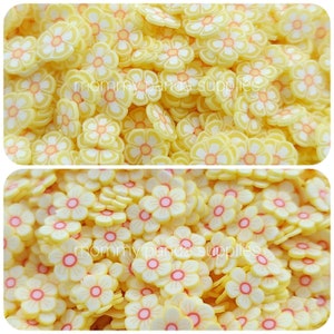 May include: A close-up image of a large quantity of yellow flower-shaped resin embellishments. The flowers have a white centre and a pink outline.