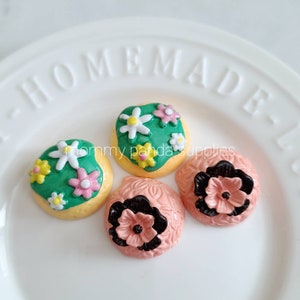May include: Three handmade cake toppers, one with green frosting and pink and white flowers, one with pink frosting and black flowers, and one with green frosting and yellow and white flowers.