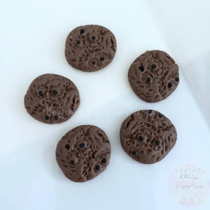 May include: Five brown chocolate chip cookie-shaped buttons with black chocolate chips. The buttons are arranged in a circle on a white surface.