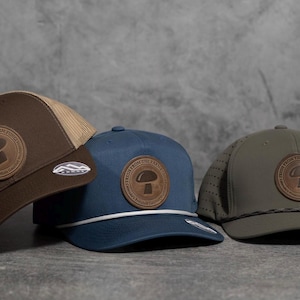 May include: Three baseball caps in brown, navy blue, and olive green. Each hat features a circular leather patch with a mushroom design and the text "FROM THE FARM". The hats have different mesh and solid panels.