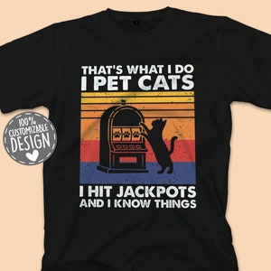 May include: Black t-shirt with a retro design featuring a cat pawing at a slot machine with the text "That's what I do I pet cats I hit jackpots and I know things".
