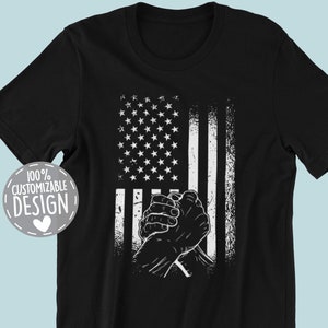 May include: Black t-shirt featuring a white graphic of clasped hands in front of a distressed American flag design. The text "100% Customizable Design" is in a white circle with a heart.