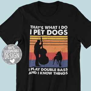 May include: Black t-shirt with a vintage-style graphic design. The text reads "That's what I do I pet dogs I play double bass and I know things". The graphic features a silhouette of a person playing a double bass and a dog.