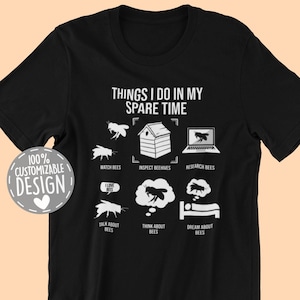 May include: Black t-shirt featuring white text and illustrations related to bees. The design includes the phrase "THINGS I DO IN MY SPARE TIME" and images of bees, a beehive, and a laptop. Additional text includes "WATCH BEES", "INSPECT BEES", "RESEARCH BEES", "TALK ABOUT BEES", "THINK ABOUT BEES", and "DREAM ABOUT BEES".