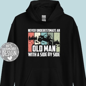 May include: Black hooded sweatshirt with the text "NEVER UNDERESTIMATE AN OLD MAN WITH A SIDE BY SIDE" in a retro design. The graphic includes a side-by-side vehicle and a forest scene. A "100% Customizable Design" badge is on the left.