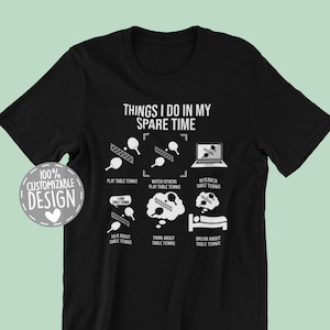 May include: Black t-shirt with a white graphic that says "Things I do in my spare time" and lists activities related to table tennis. The graphic includes a table tennis paddle, a ball, a laptop, and a bed. The text "100% Customizable Design" is in a white circle with a heart in the center.
