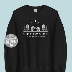 May include: Black sweatshirt with a white design of a side-by-side vehicle driving through a forest at night. The text "Side by Side is How We Ride" is printed below the vehicle.