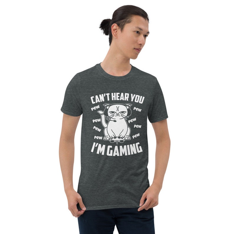 May include: A dark heather grey t-shirt with a white graphic of an angry cat wearing headphones and holding a video game controller. The text "Can't hear you I'm gaming" is printed on the shirt, surrounded by the word "pew" repeated multiple times.
