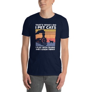 May include: A navy blue t-shirt with a vintage-style graphic of a train and a cat. The text on the shirt reads "That's what I do I pet cats I play with trains and I know things."