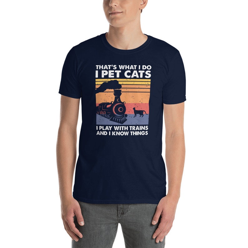 May include: A navy blue t-shirt with a vintage-style graphic of a train and a cat. The text on the shirt reads "That's what I do I pet cats I play with trains and I know things."