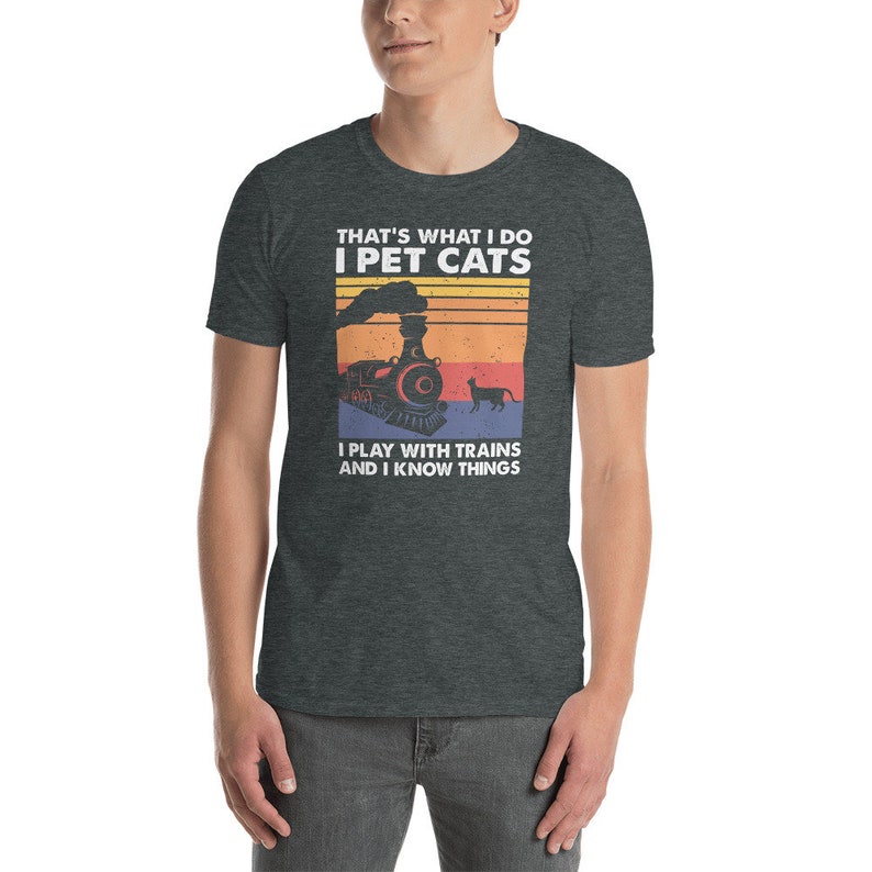 May include: A dark heather grey t-shirt with a vintage-style graphic of a train and a cat. The text on the shirt reads "That's what I do I pet cats I play with trains and I know things."