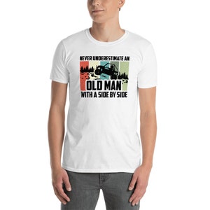 以下が含まれることがあります： 白地に、ヴィンテージスタイルのグラフィックがプリントされたTシャツ。グラフィックには、「Never underestimate an old man with a side by side」というテキストと、森の中をサイドバイサイドの乗り物に乗っている人のシルエットが描かれています。