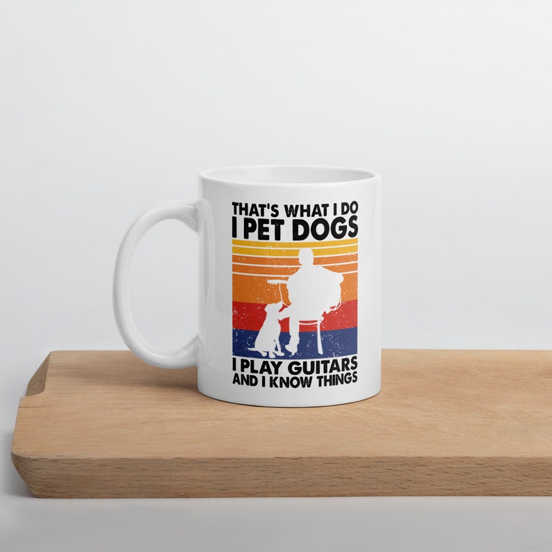 Puede incluir: Taza de cer&aacute;mica blanca con un dise&ntilde;o retro que presenta una silueta de una persona tocando una guitarra con un perro sentado a su lado. El texto dice "That's what I do I pet dogs I play guitars and I know things".