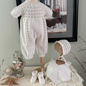 May include: A white crochet baby outfit set, including a romper, bonnet, bib, and shoes. The romper is displayed on a wooden stand. The set is handmade with intricate openwork details, perfect for a newborn. The shoes are approximately 4 inches long.