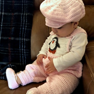 May include: A baby wearing a pink crocheted outfit with a penguin design on the overalls. The outfit includes a matching hat, pants, and booties.