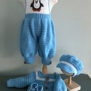 Puede incluir: Un conjunto de ropa de bebé de punto azul con un diseño de pingüino en el mono, una chaqueta a juego, un gorro y botines.