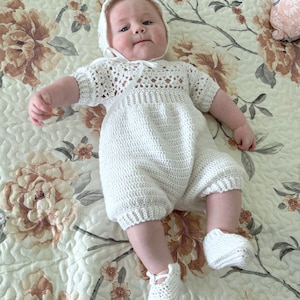 May include: A white crocheted baby outfit with a matching bonnet and booties. The outfit features a delicate lace pattern and a bow at the neckline.