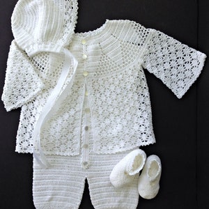 May include: A white crocheted baby outfit set, including a bonnet, cardigan, pants, and booties. The set is made with delicate lace patterns and features button closures.