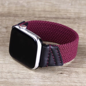 May include: A silver smartwatch with a black screen and a red digital crown, attached to a dark burgundy braided elastic watch band. The band features black leather connectors on both sides, secured with visible dark red stitching detail.