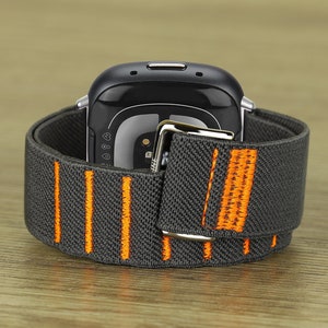 Puede incluir: Una correa de reloj el&aacute;stica gris y naranja con una hebilla plateada. La correa est&aacute; dise&ntilde;ada para un reloj inteligente con una esfera redonda.