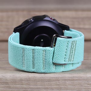 Pode incluir: Uma pulseira de relógio elástica azul claro com uma fivela prateada. A pulseira é projetada para um smartwatch com uma caixa preta.