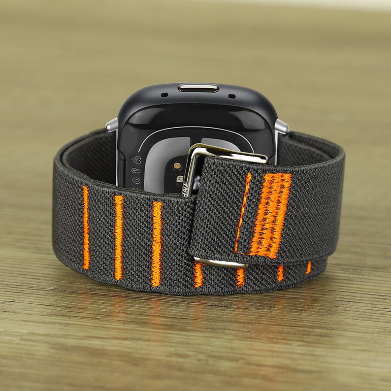 Puede incluir: Una correa de reloj el&aacute;stica gris y naranja con una hebilla plateada. La correa est&aacute; dise&ntilde;ada para un reloj inteligente con una esfera redonda.