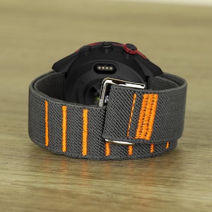 Könnte beinhalten: Eine schwarze Smartwatch mit einem grauen und orangefarbenen Stoffband. Die Uhr hat einen roten Akzent auf der Lünette und eine silberne Schnalle.