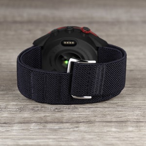 May include: Black and red smartwatch with a black elastic band. The watch has a round face with a black bezel and a silver buckle.