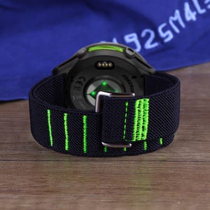 Könnte beinhalten: Ein schwarzes und grünes elastisches Uhrenarmband mit einer silbernen Schnalle ist an einer schwarzen Smartwatch befestigt. Das Armband hat grüne vertikale Streifen. Die Uhr liegt auf einer Holzoberfläche.