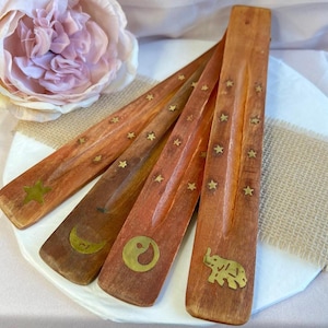 May include: Four wooden incense holders with brass star, moon, yin-yang, and elephant designs. The holders are various shades of brown and red-brown. The holders are on a white plate with a burlap and floral background.