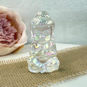 May include: A small, iridescent Buddha statue in a seated meditation pose. The figurine has a pearlescent finish with hints of pink, blue, and yellow. The statue is on a burlap surface, with a soft pink flower in the background.