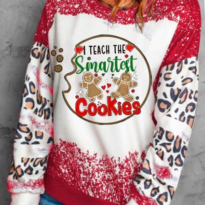 May include: A white and red sweatshirt with the text "I Teach The Smartest Cookies." The design features gingerbread people and hearts. The sleeves have a leopard print pattern.