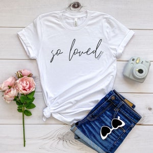 May include: White t-shirt with the text "so loved" in black cursive script and the text "John 3:16" in smaller black font below. The shirt is laid out on a light wood surface with a pair of blue denim shorts, a pair of sunglasses, and a pink flower bouquet.