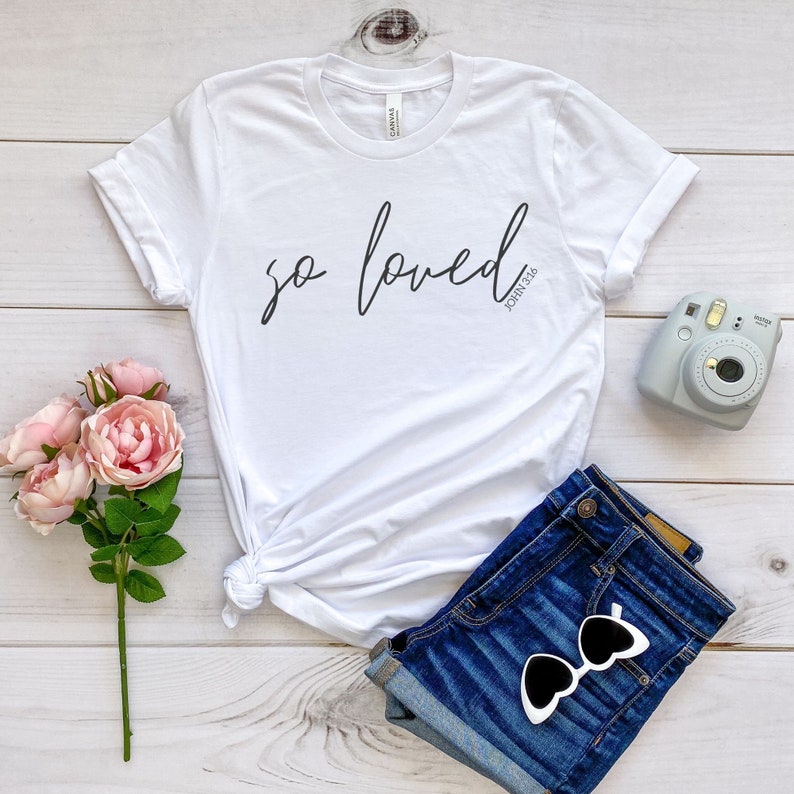 May include: White t-shirt with the text "so loved" in black cursive script and the text "John 3:16" in smaller black font below. The shirt is laid out on a light wood surface with a pair of blue denim shorts, a pair of sunglasses, and a pink flower bouquet.