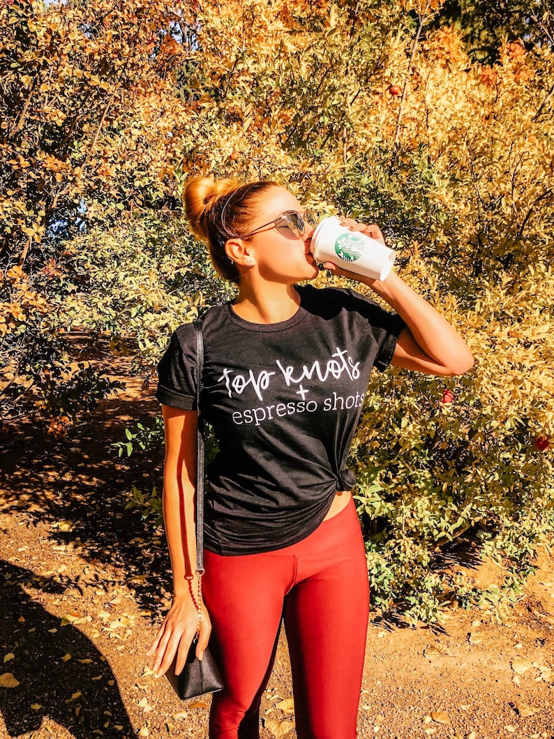 May include: A woman wearing red leggings and a black t-shirt with the text "Top Knots + Espresso Shots" is drinking from a white coffee cup.
