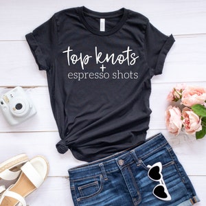 May include: Black t-shirt with white text that reads "Top Knots + Espresso Shots". The shirt is on a white wooden surface with a pair of white sandals, a white instant camera, and a bouquet of pink flowers.