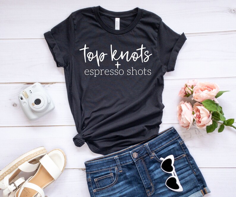 May include: Black t-shirt with white text that reads "Top Knots + Espresso Shots". The shirt is on a white wooden surface with a pair of white sandals, a white instant camera, and a bouquet of pink flowers.
