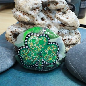 May include: A painted rock with a green shamrock design. The shamrock is surrounded by white dots and a black outline. The rock is sitting on a gray rock.