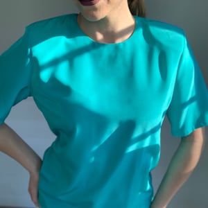 May include: A turquoise short-sleeved blouse with a round neckline. The blouse is made of a silky material and has a relaxed fit.