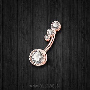 May include: Rose gold belly button ring with four clear, round, sparkling gemstones. The largest gem is at the base, with three smaller gems ascending. The jewelry is set against a dark wood grain background. The text "ANMOL JEWELS" is at the bottom.
