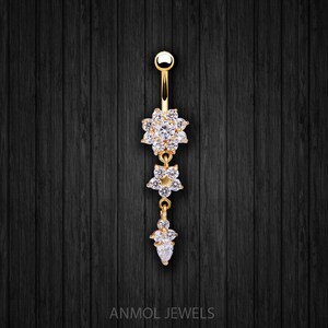 May include: Gold-toned belly button ring with three floral-shaped charms, each encrusted with clear crystals. The top charm is a large flower, followed by a smaller flower, and a teardrop-shaped charm at the bottom. The text "ANMOL JEWELS" is at the bottom.