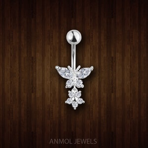 May include: A silver belly button ring featuring a butterfly and flower design. The butterfly has marquise-cut stones for wings and round stones for the body. A cluster of round stones forms the flower. The brand name "ANMOL JEWELS" is at the bottom.