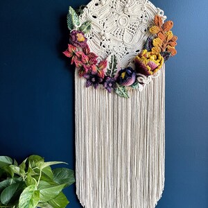 May include: A white macrame wall hanging with a circular design and a fringe border. The hanging is decorated with crocheted flowers in shades of red, purple, yellow, and green.
